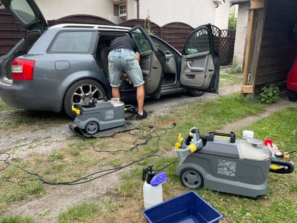 Mężczyzna w dżinsowych szortach czyści wnętrze szarego kombi za pomocą profesjonalnego odkurzacza piorącego, drugi odkurzacz i akcesoria leżą na trawie obok samochodu.