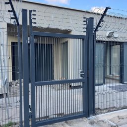 ZEGGER TECH SPÓŁKA Z OGRANICZONĄ ODPOWIEDZIALNOŚCIĄ - Szara metalowa brama z pionowymi prętami i drutem kolczastym na górze, zamontowana w ogrodzeniu panelowym, widok na budynek w tle.