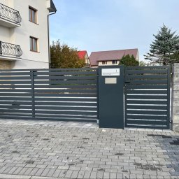ZEGGER TECH SPÓŁKA Z OGRANICZONĄ ODPOWIEDZIALNOŚCIĄ - Nowoczesna, przesuwna brama wjazdowa w kolorze grafitowym z poziomymi panelami, zintegrowana ze skrzynką na listy i tabliczką z numerem domu 'Różana 1', osadzona na słupkach z szarego betonu...