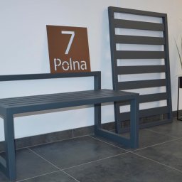 ZEGGER TECH SPÓŁKA Z OGRANICZONĄ ODPOWIEDZIALNOŚCIĄ - Nowoczesna ławka ogrodowa z metalu w kolorze grafitowym, obok panelowa przesłona, adres '7 Polna' na brązowej tabliczce, minimalistyczna donica z trawą pampasową w tle.