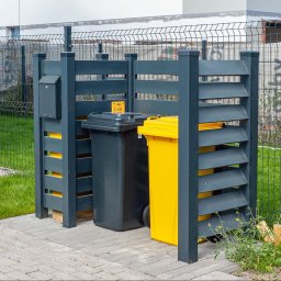 ZEGGER TECH SPÓŁKA Z OGRANICZONĄ ODPOWIEDZIALNOŚCIĄ - Zabudowa śmietnikowa z szarego drewna z lamelami, ukrywająca dwa pojemniki na śmieci: czarny i żółty, na tle panelowego ogrodzenia.