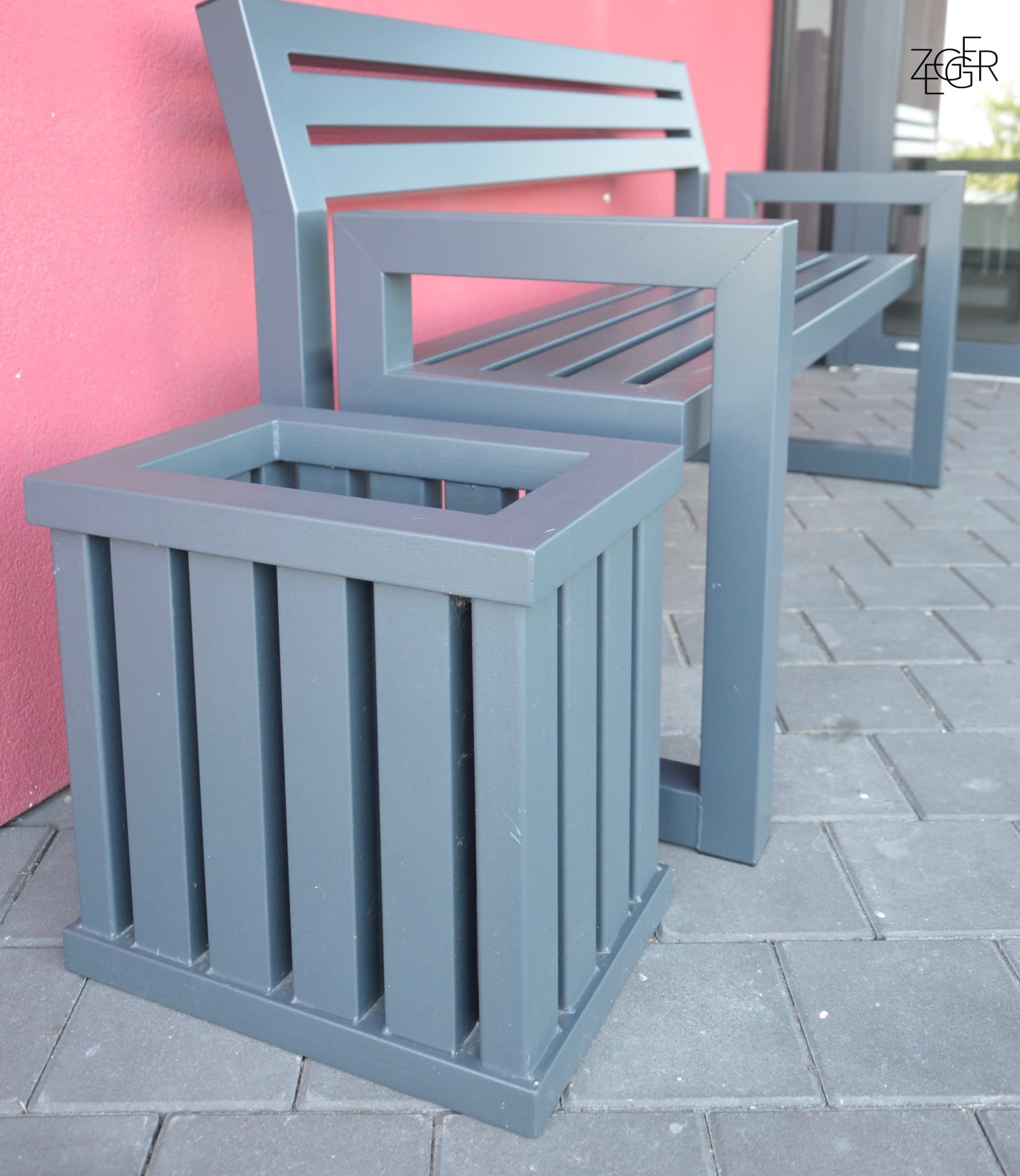 Szara metalowa ławka z oparciem i podłokietnikami, obok donica z tego samego materiału, na szarej kostce brukowej i tle różowej ściany.