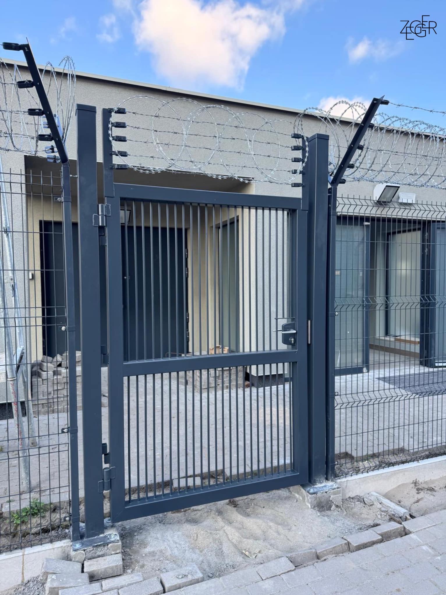 Szara metalowa brama z pionowymi prętami i drutem kolczastym na górze, zamontowana w ogrodzeniu panelowym, widok na budynek w tle.
