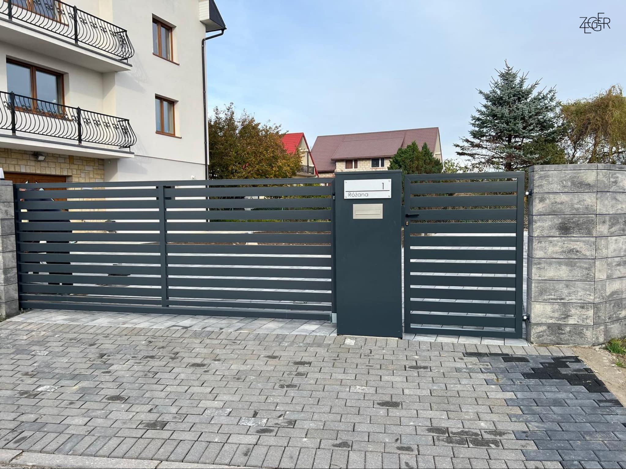 Nowoczesna, przesuwna brama wjazdowa w kolorze grafitowym z poziomymi panelami, zintegrowana ze skrzynką na listy i tabliczką z numerem domu 'Różana 1', osadzona na słupkach z szarego betonu...
