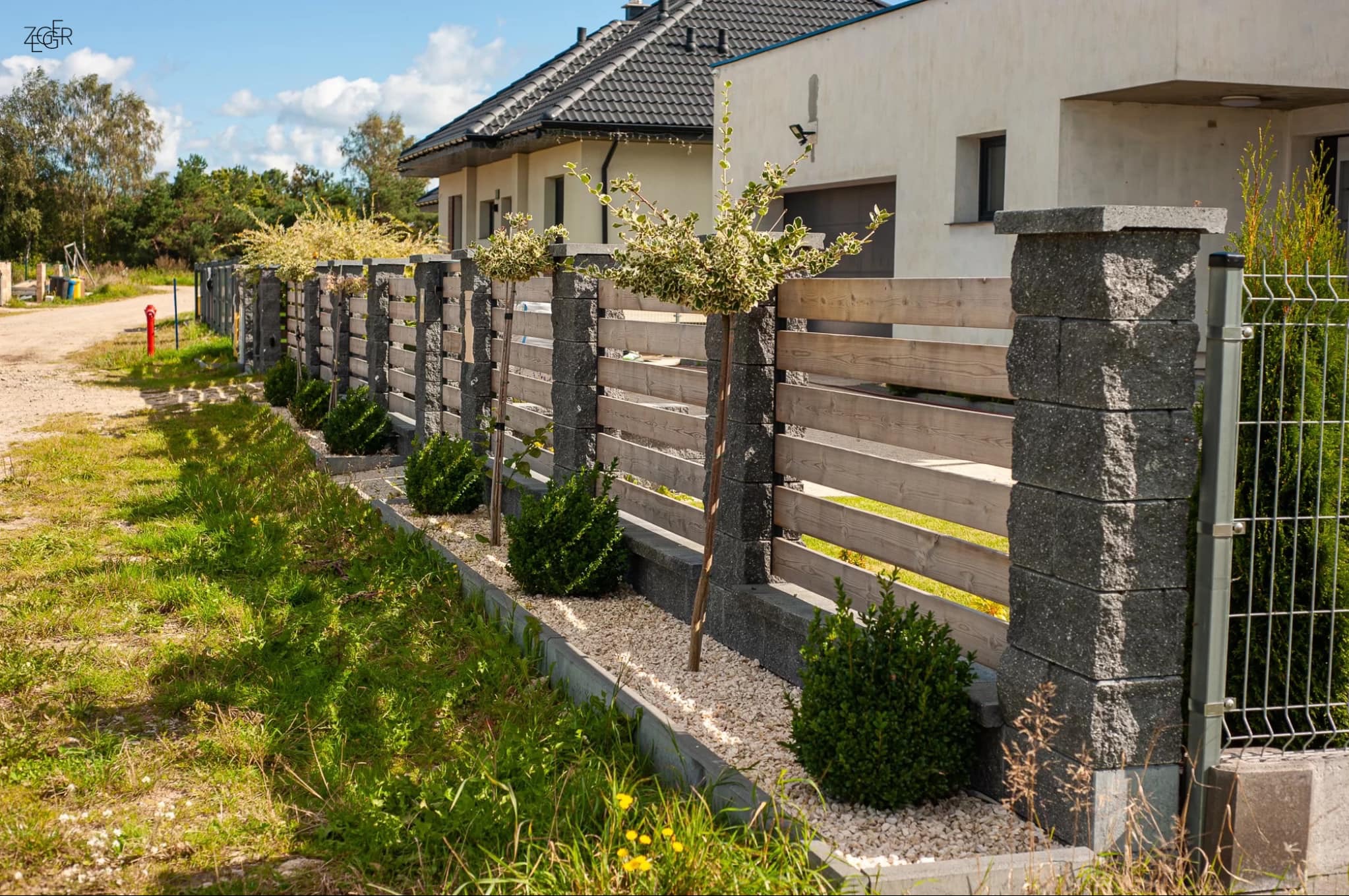 Nowoczesne ogrodzenie z betonowych słupków i drewnianych paneli, ozdobione niskimi krzewami i jasnym żwirem, widoczne na tle domu z ciemnym dachem.