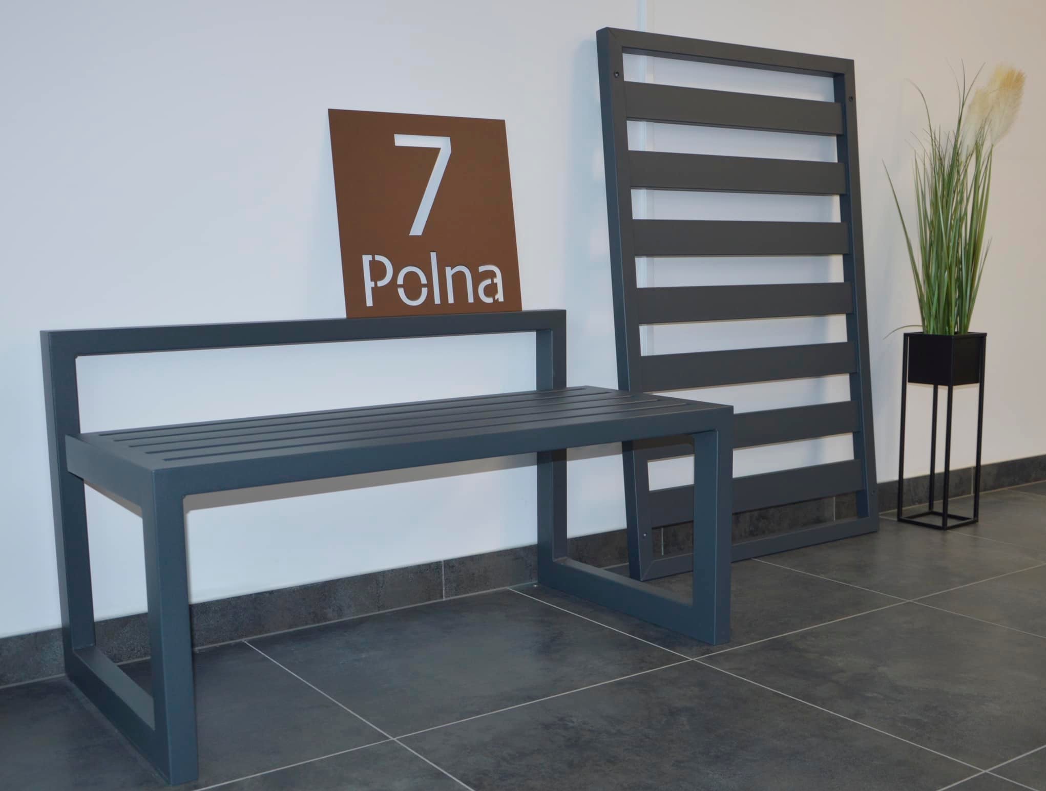 Nowoczesna ławka ogrodowa z metalu w kolorze grafitowym, obok panelowa przesłona, adres '7 Polna' na brązowej tabliczce, minimalistyczna donica z trawą pampasową w tle.