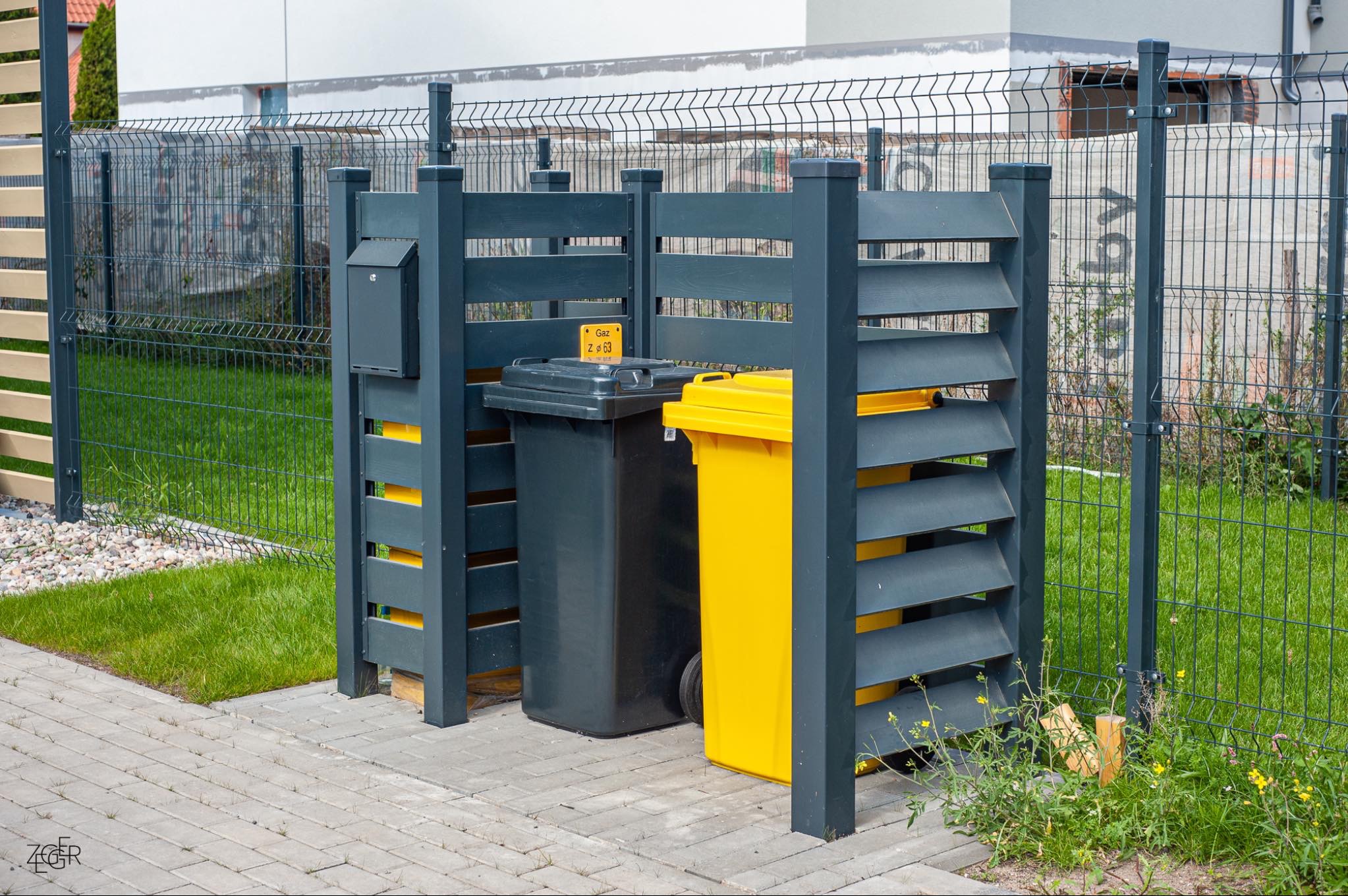 Zabudowa śmietnikowa z szarego drewna z lamelami, ukrywająca dwa pojemniki na śmieci: czarny i żółty, na tle panelowego ogrodzenia.