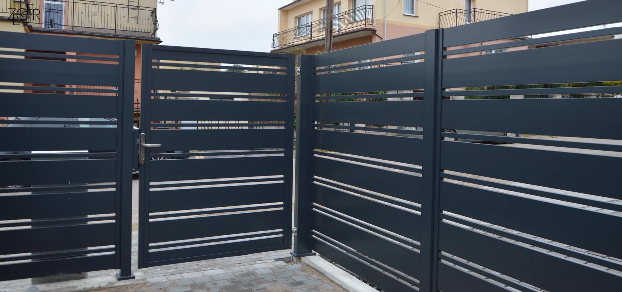 Nowoczesna, grafitowa brama wjazdowa z poziomymi, jasnymi prześwitami, częściowo otwarta, z widocznym fragmentem posesji i domu w tle.