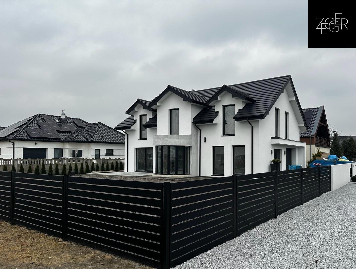 Nowoczesny dom jednorodzinny z czarną, poziomą zabudową ogrodzeniową i szarym żwirem na podjeździe, widok z zewnątrz w pochmurny dzień.