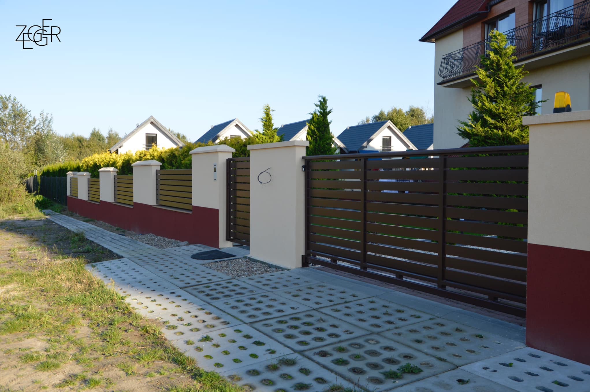 Nowoczesne ogrodzenie posesji z poziomych, brązowych paneli z bramą przesuwną, osadzone na słupkach z jasnego betonu z czerwoną podmurówką, widok na podjazd z ażurowych płyt betonowych.
