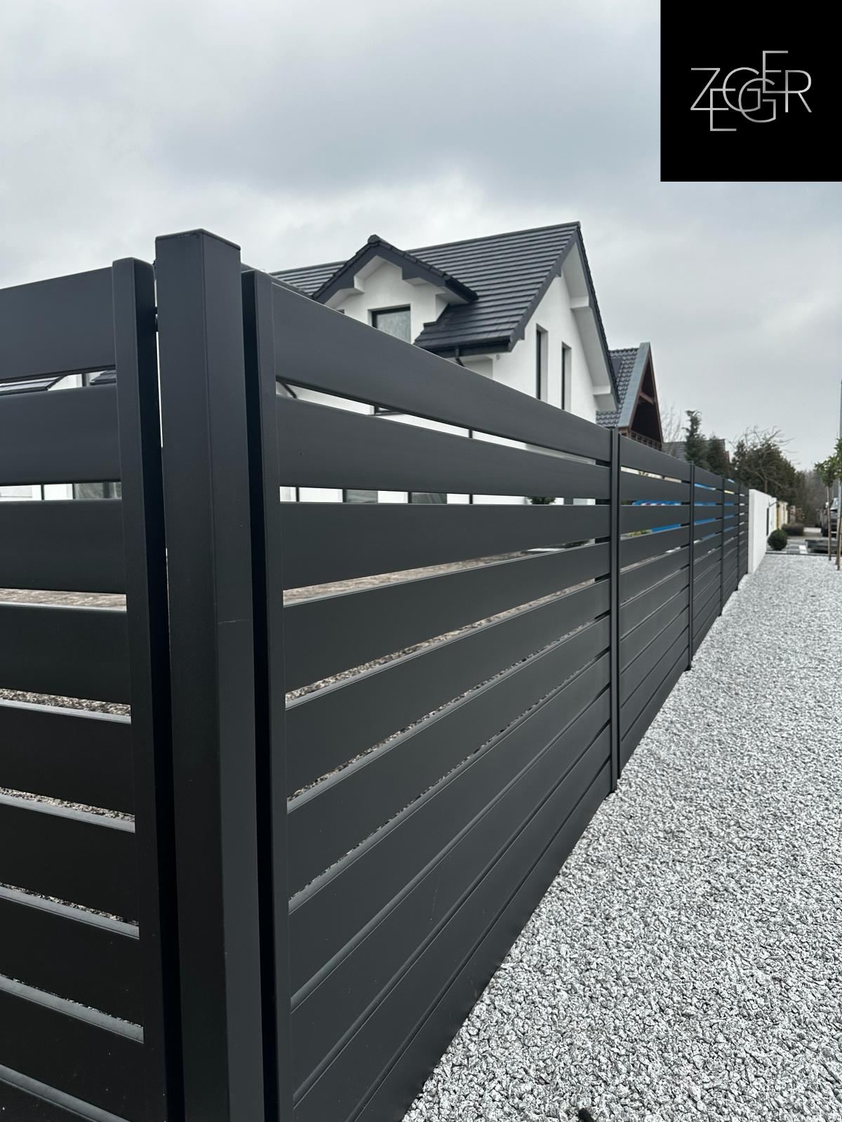 Nowoczesne, poziome ogrodzenie z metalowych profili w kolorze grafitowym, wykończone drobnym, białym żwirem, z widocznym logo firmy Zeger w prawym górnym rogu i fragmentem białego domu w tle.