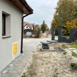 JOP-KOP Michał Pietras - Prace przygotowawcze do ułożenia kostki brukowej: robotnik wyrównuje teren podłoża, widoczne palety z kostką, ułożone fragmenty obrzeży i budynek z żółtą skrzynką gazową na ścianie.