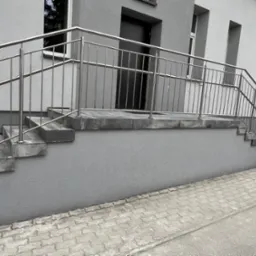 Zewnętrzne schody wejściowe do budynku wyłożone szarą kostką, z metalową balustradą, prowadzące do ciemnych drzwi wejściowych.