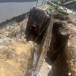 JOP-KOP Michał Pietras - Wykop fundamentowy zbrojony stalową konstrukcją przy budynku, widoczny pracownik w tle i pomarańczowy kabel na ziemi.