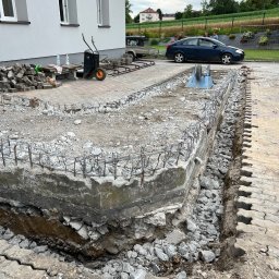JOP-KOP Michał Pietras - Przygotowanie podłoża pod kostkę brukową: widoczny wykop z betonową obudową wzmocnioną stalowymi prętami, częściowo wypełniony gruzem, z ułożoną obok kostką brukową. W tle budynek, taczki...