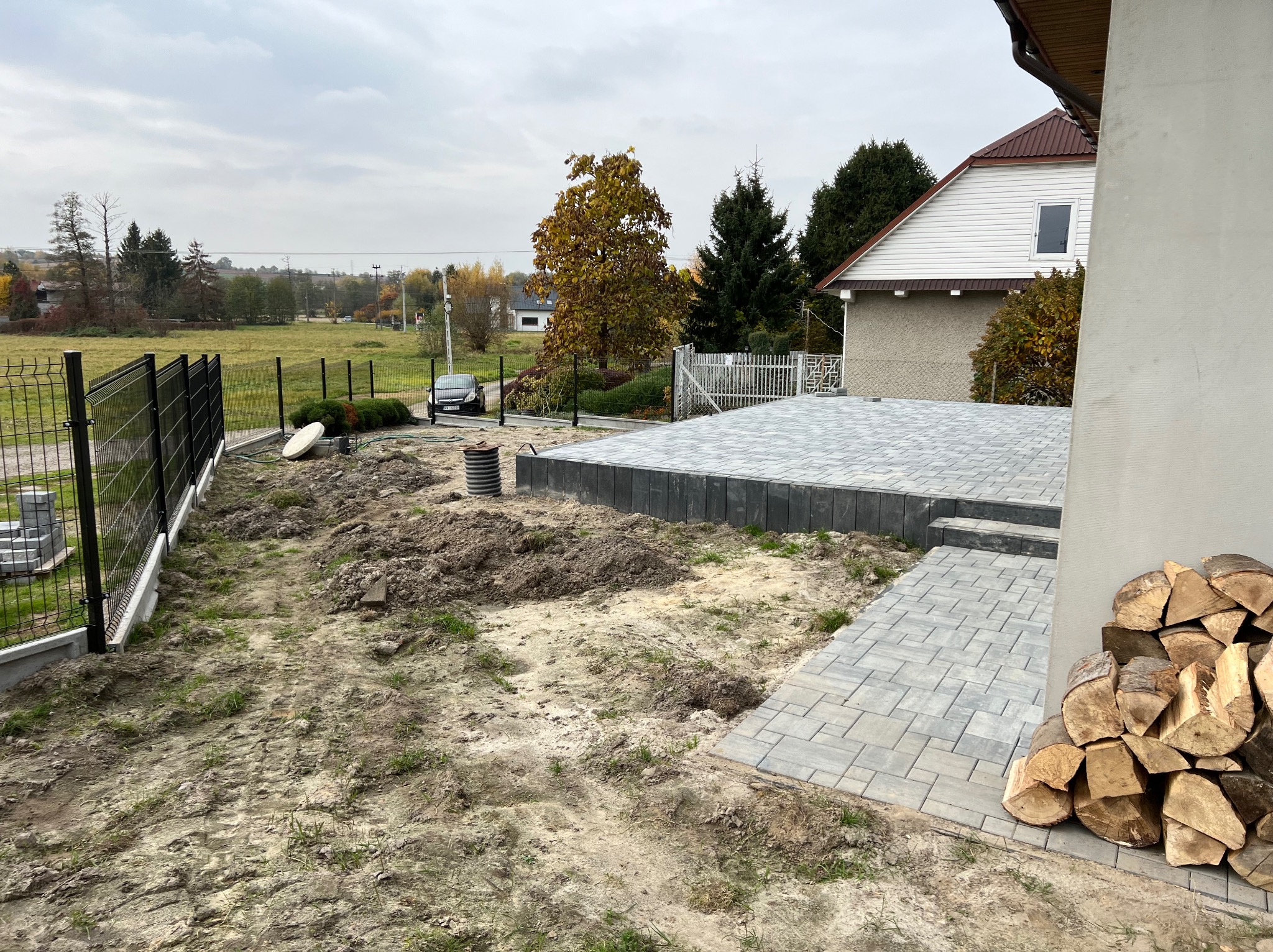 Przygotowanie terenu pod kostkę brukową na posesji: częściowo ułożona nawierzchnia z kostki, pryzmy ziemi, widoczny drenaż, czarne ogrodzenie panelowe i sterta drewna kominkowego pod ścianą domu.