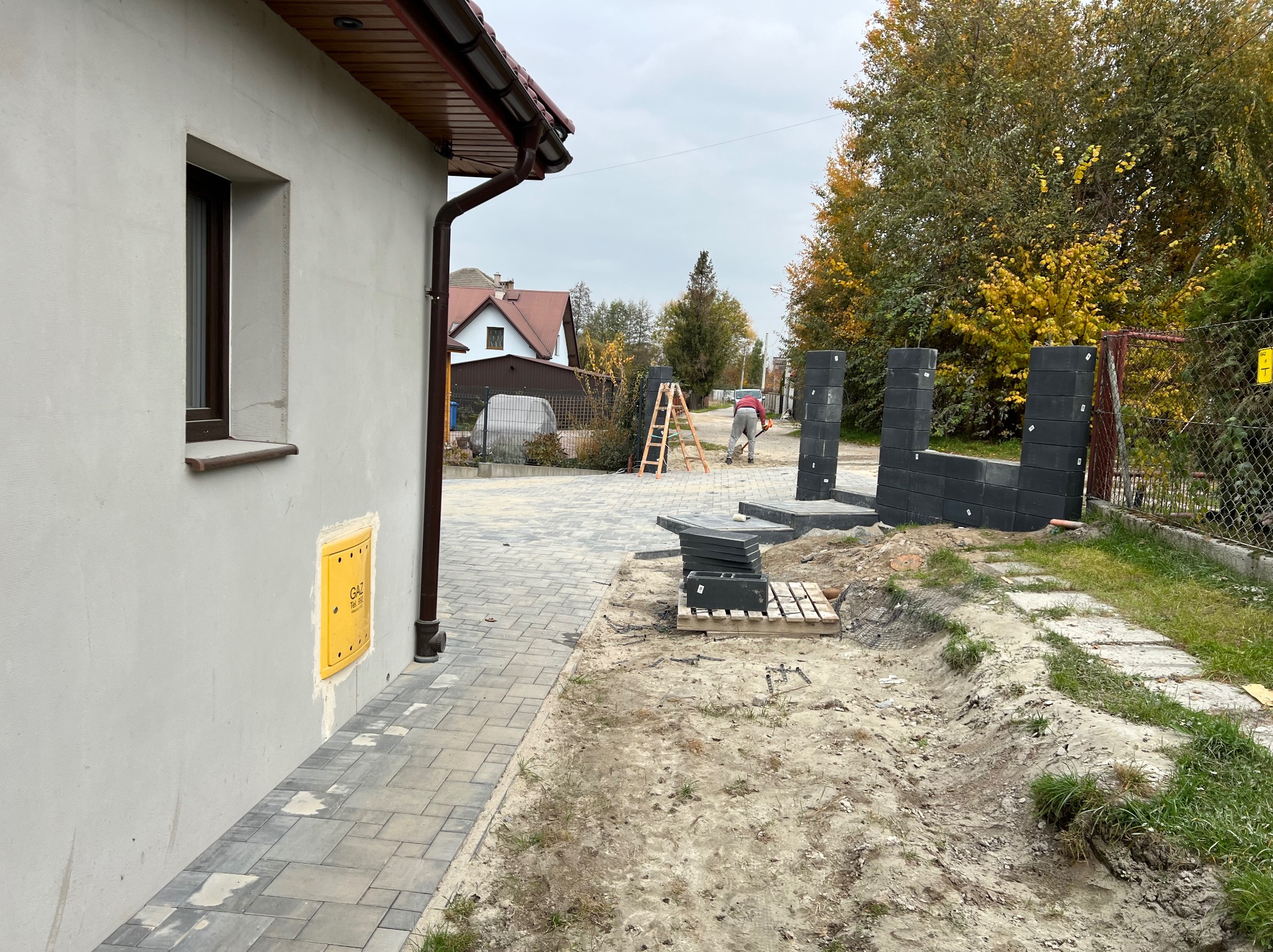 Prace przygotowawcze do ułożenia kostki brukowej: robotnik wyrównuje teren podłoża, widoczne palety z kostką, ułożone fragmenty obrzeży i budynek z żółtą skrzynką gazową na ścianie.