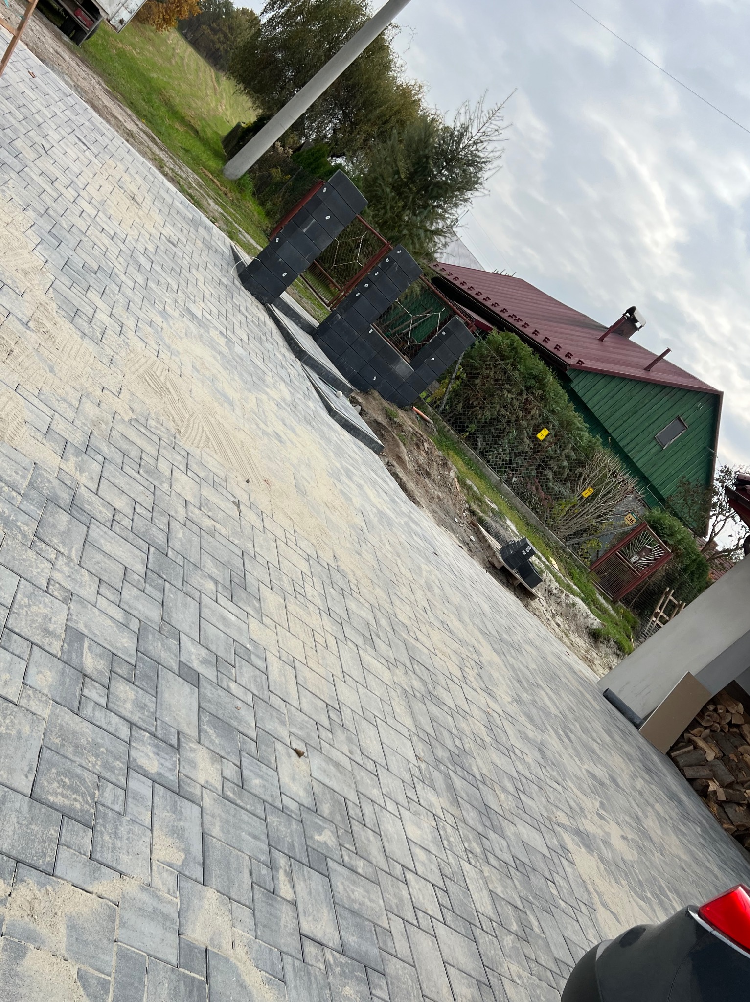 Świeżo ułożona, szara kostka brukowa na podjeździe, z widocznymi śladami piasku i przygotowanymi elementami do dalszej pracy, na tle zielonego domu z czerwoną dachówką.