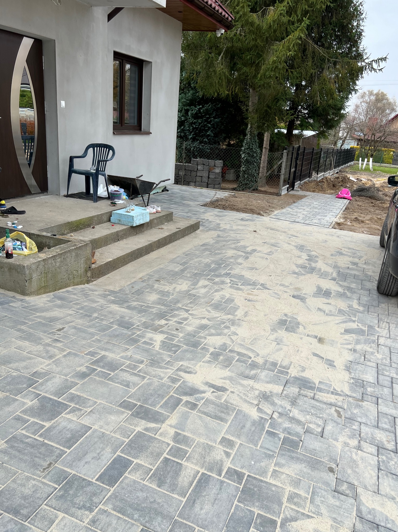 Ułożona kostka brukowa w Sędziszowicach, widok z góry na szare, różnej wielkości kostki. Na schodach przed domem leżą narzędzia i przedmioty. W tle widać częściowo ogrodzenie.