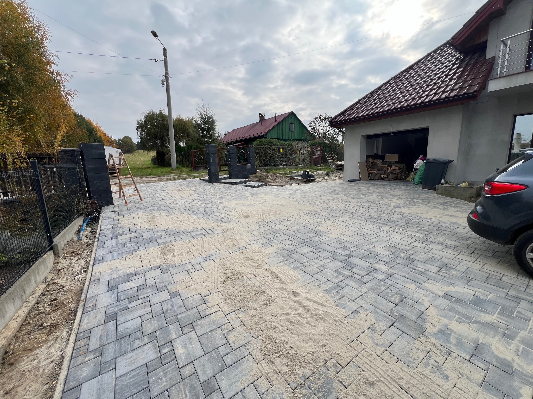 Ułożona kostka brukowa na podjeździe posesji z widocznymi śladami opon, częściowo zasypana piaskiem, obok zaparkowany samochód, w tle dom z garażem i ogród.