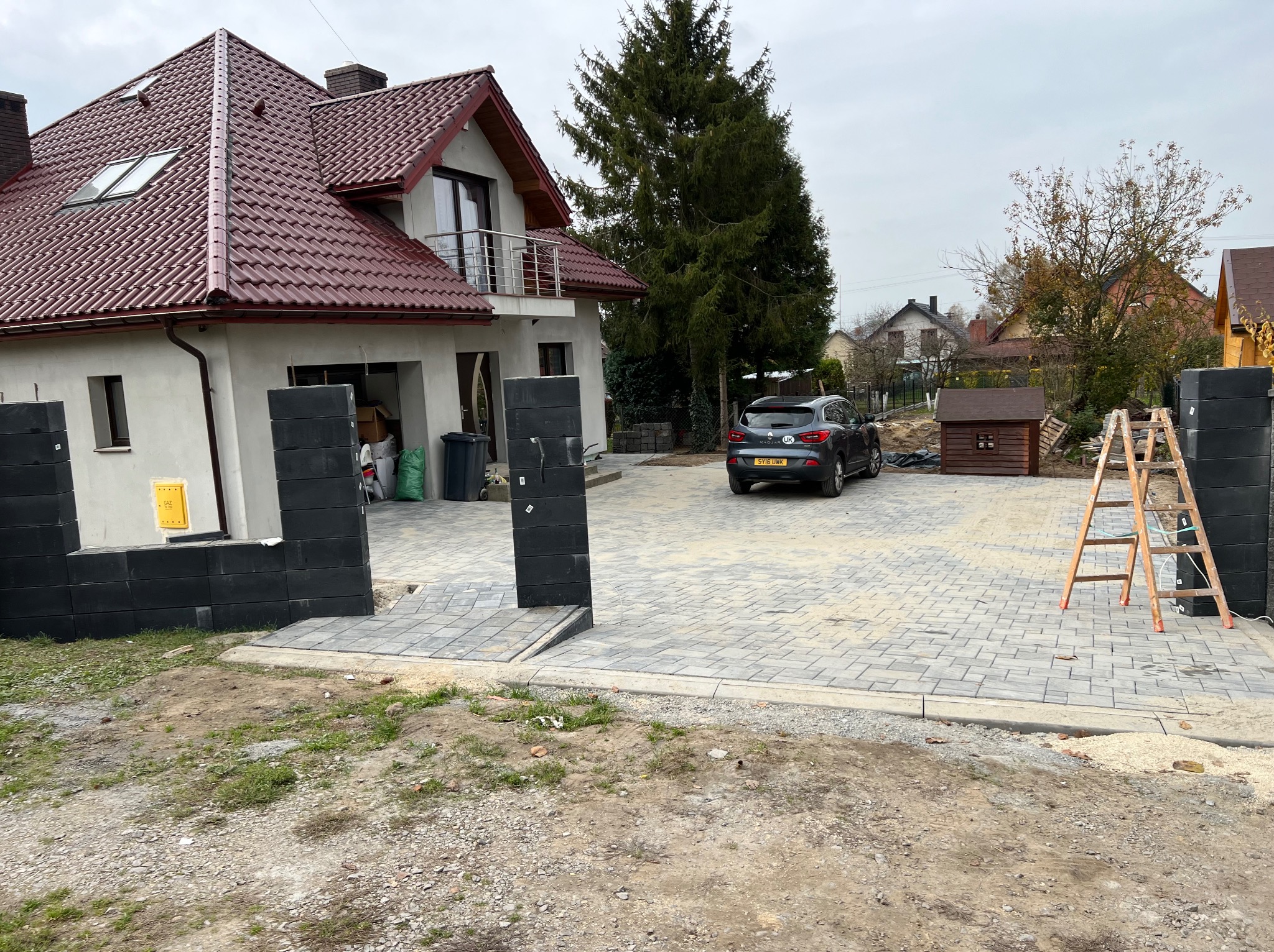 Ułożona szara kostka brukowa na podjeździe przed domem z czerwonym dachem, widoczne czarne słupki ogrodzeniowe, drabina i zaparkowany samochód.