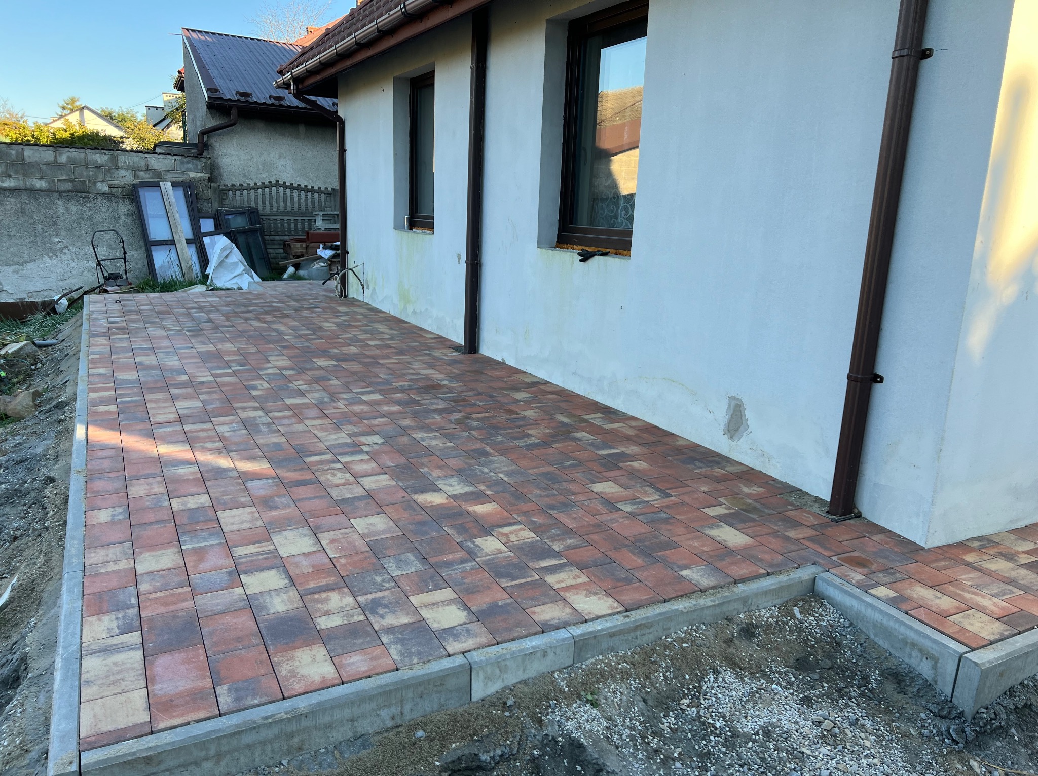 Nowo ułożona kostka brukowa w odcieniach czerwieni i brązu, obramowana betonowym krawężnikiem, przylegająca do ściany budynku z brązowymi rynnami.