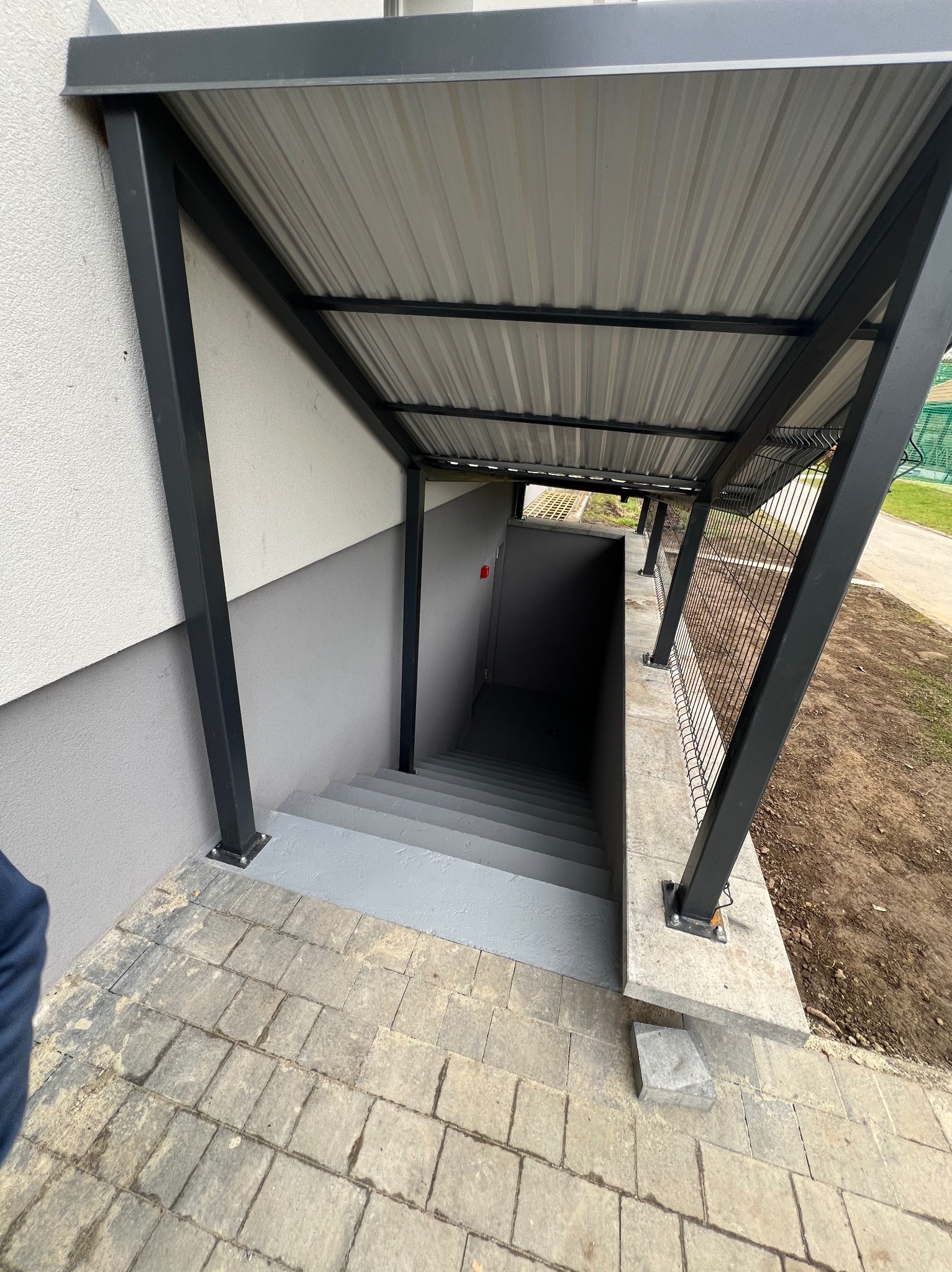 Zadaszone schody do piwnicy z szarą kostką brukową i metalową konstrukcją z siatką po prawej stronie.