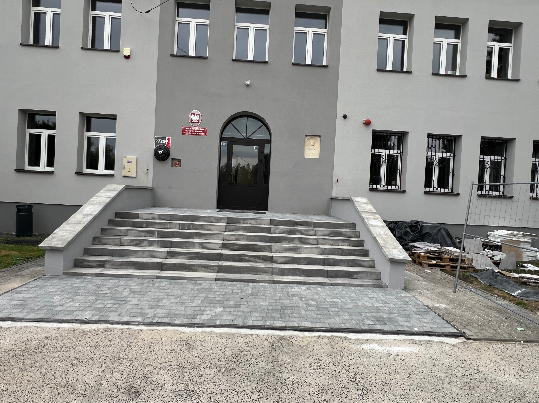 Nowo ułożona kostka brukowa przed budynkiem szkoły z szarymi schodami prowadzącymi do wejścia, widoczne materiały budowlane w tle za ogrodzeniem.