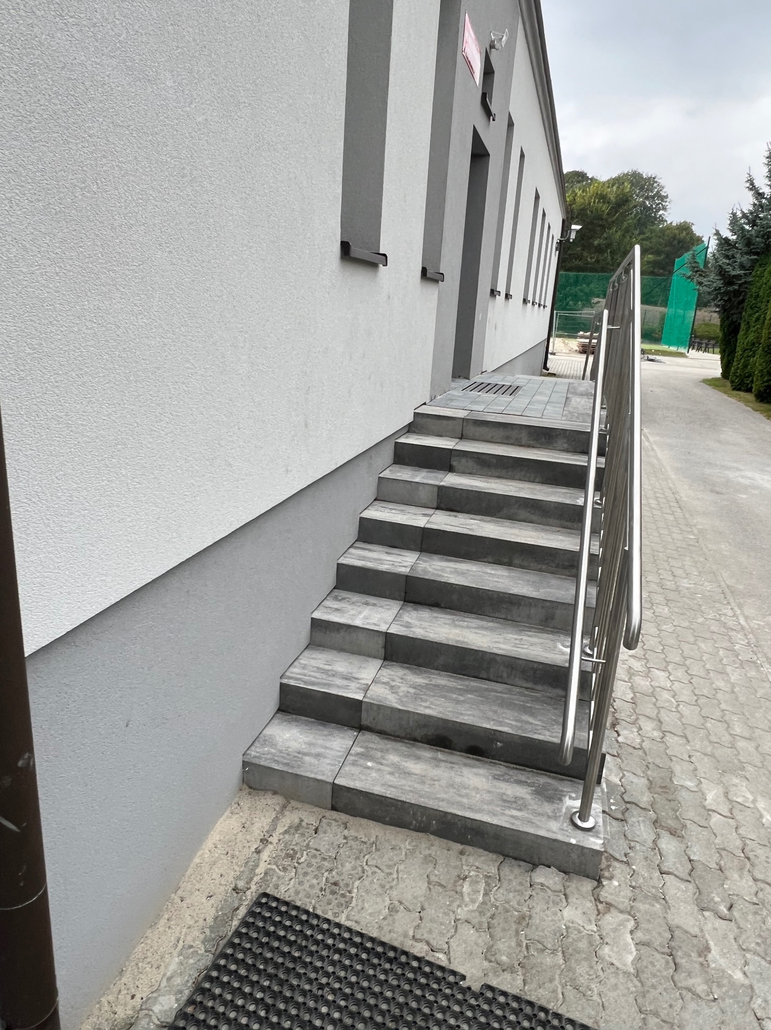Schody zewnętrzne wyłożone szarą kostką brukową prowadzące do budynku z metalową barierką, widok z boku, obok chodnik z kostki.