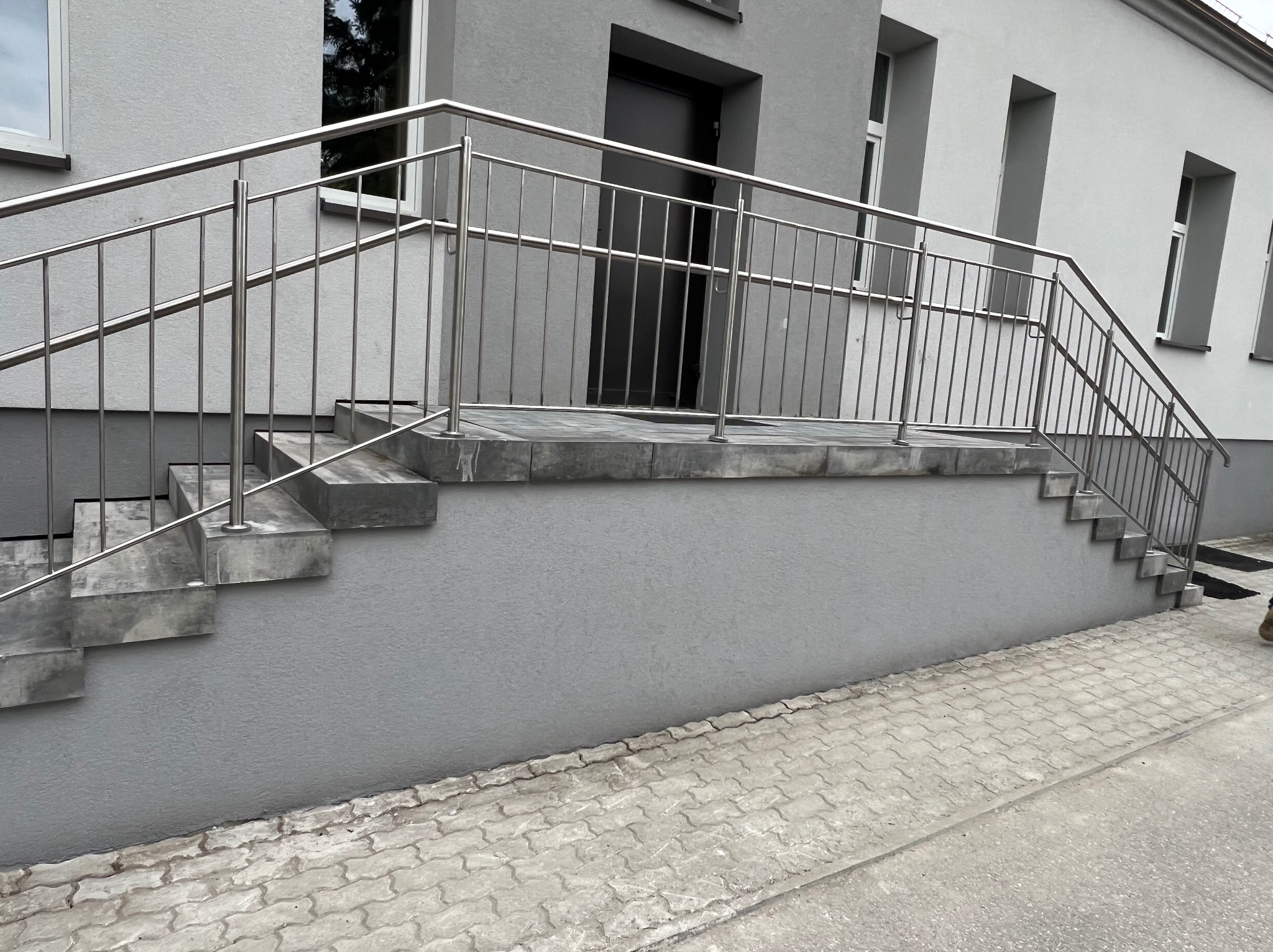 Zewnętrzne schody wejściowe do budynku wyłożone szarą kostką, z metalową balustradą, prowadzące do ciemnych drzwi wejściowych.