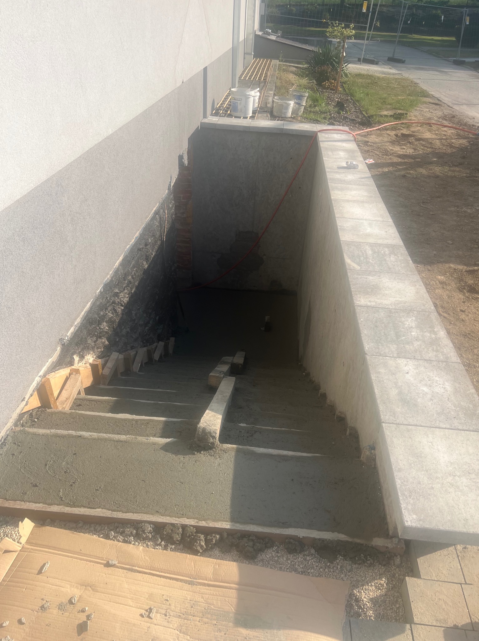 Betonowe schody zewnętrzne w trakcie budowy, z drewnianymi szalunkami i częściowo ułożonymi płytami chodnikowymi, prowadzące do ciemnego wejścia.