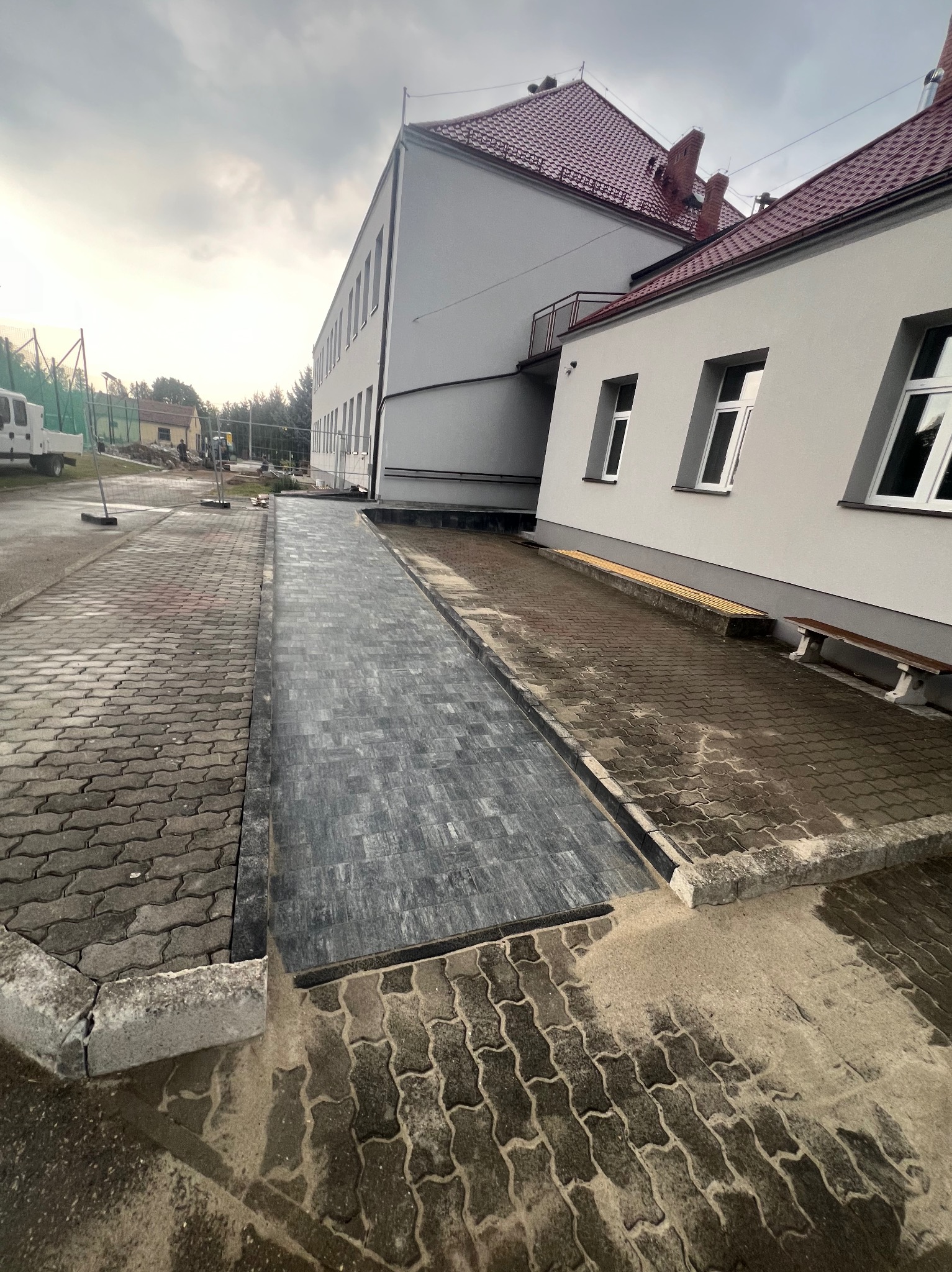 Ułożona kostka brukowa wzdłuż budynku z czerwonym dachem, z widoczną różnicą w kolorze i wzorze kostki na nowym odcinku, częściowo mokra powierzchnia, pochmurne niebo.