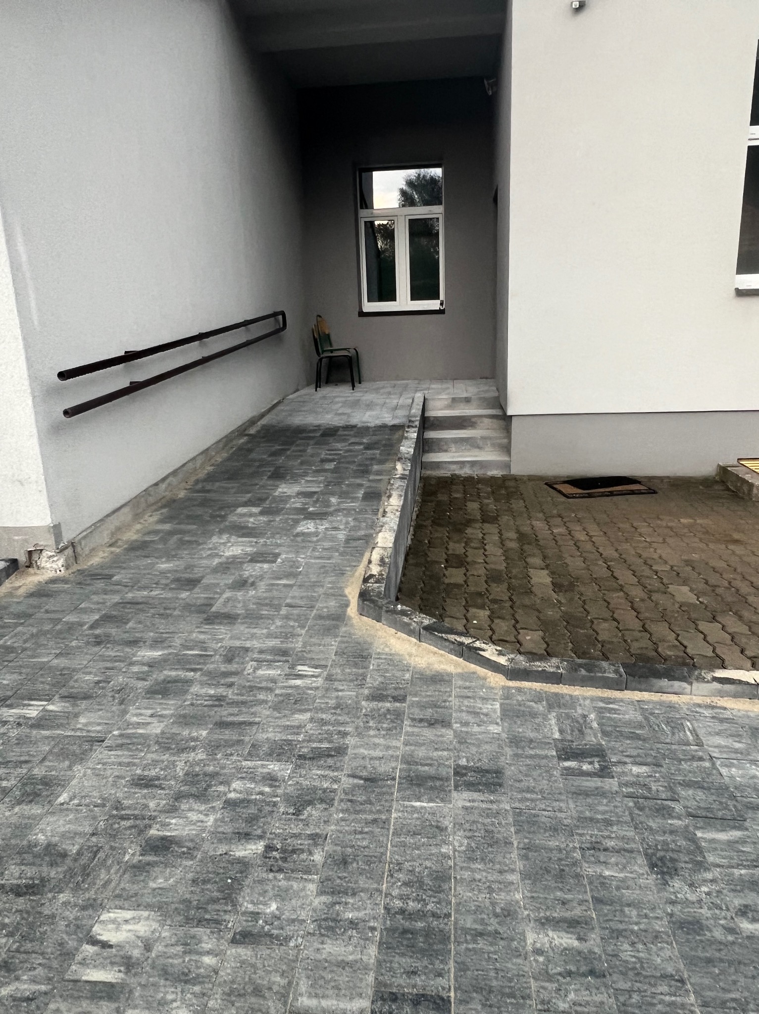 Ułożona kostka brukowa w kolorze szaro-grafitowym, tworząca podjazd z rampą dla wózków inwalidzkich, prowadząca do wejścia budynku z widocznym oknem i krzesłem w głębi.