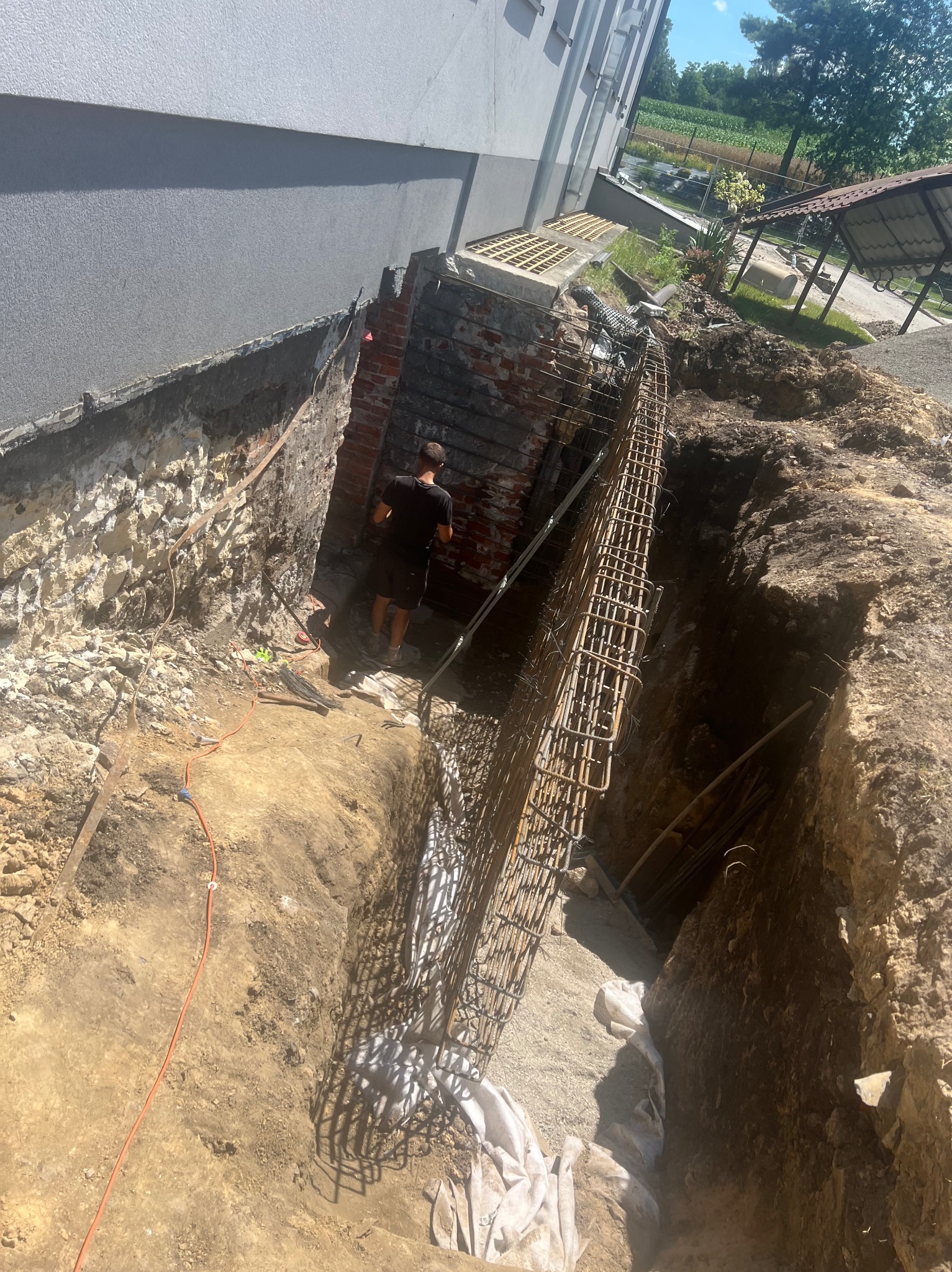 Wykop fundamentowy zbrojony stalową konstrukcją przy budynku, widoczny pracownik w tle i pomarańczowy kabel na ziemi.