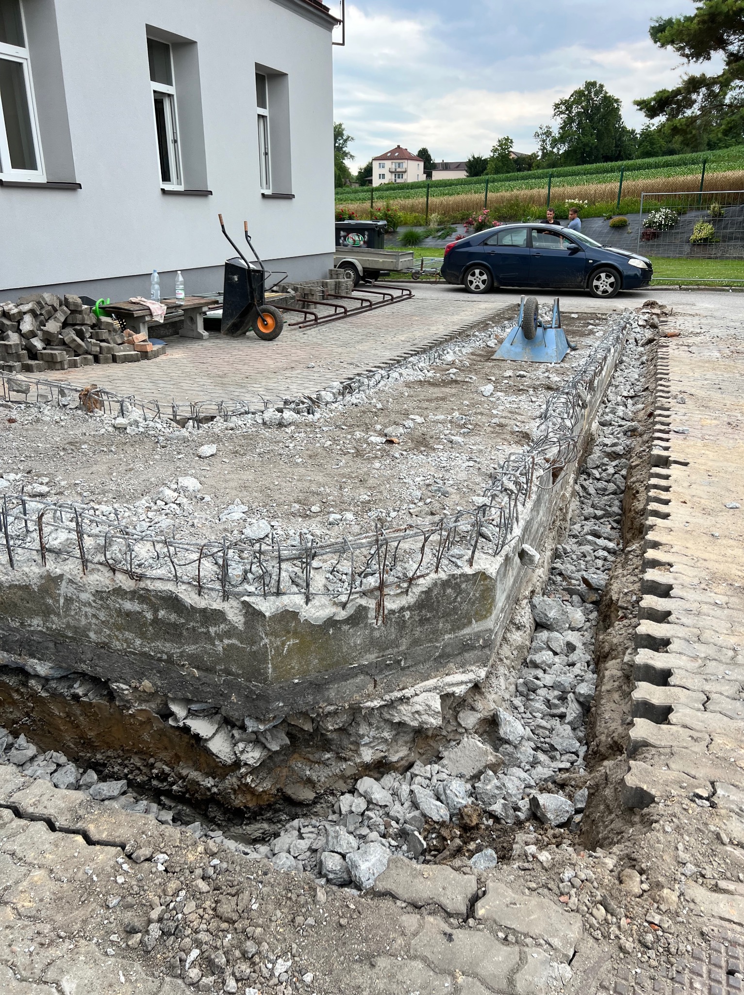 Przygotowanie podłoża pod kostkę brukową: widoczny wykop z betonową obudową wzmocnioną stalowymi prętami, częściowo wypełniony gruzem, z ułożoną obok kostką brukową. W tle budynek, taczki...