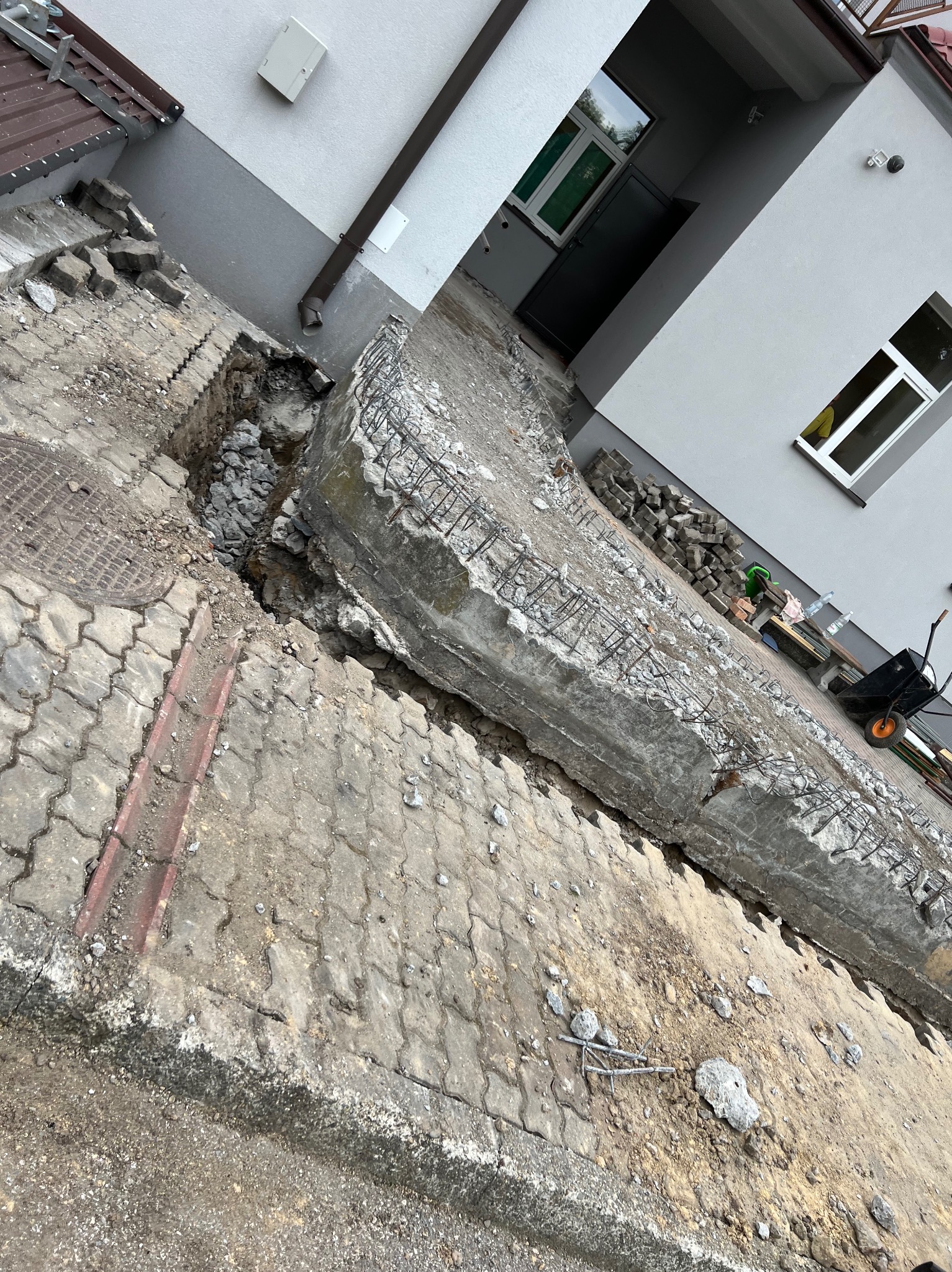 Fragment rozkopanego chodnika z kostki brukowej, odsłonięty wykop z kamieniami i fragmentem zbrojonego betonu, w tle ściana budynku z oknem i rynną.