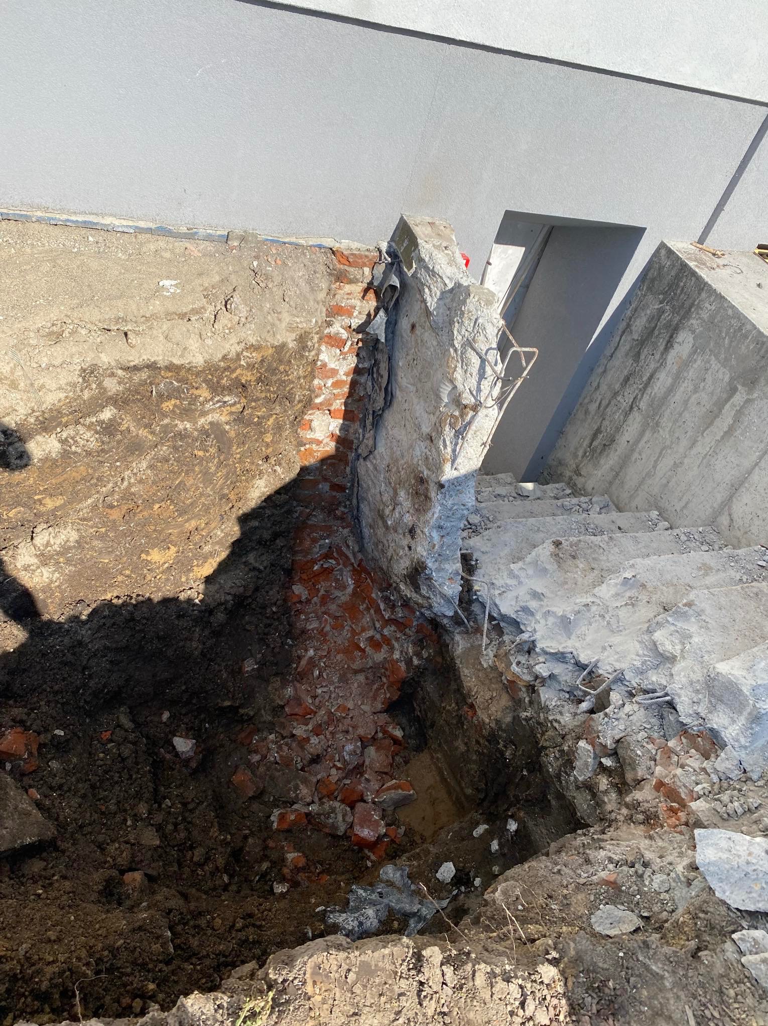 Odkopana ściana fundamentowa z cegły, częściowo zniszczone betonowe schody i wykopana ziemia wokół, widoczne pręty zbrojeniowe.