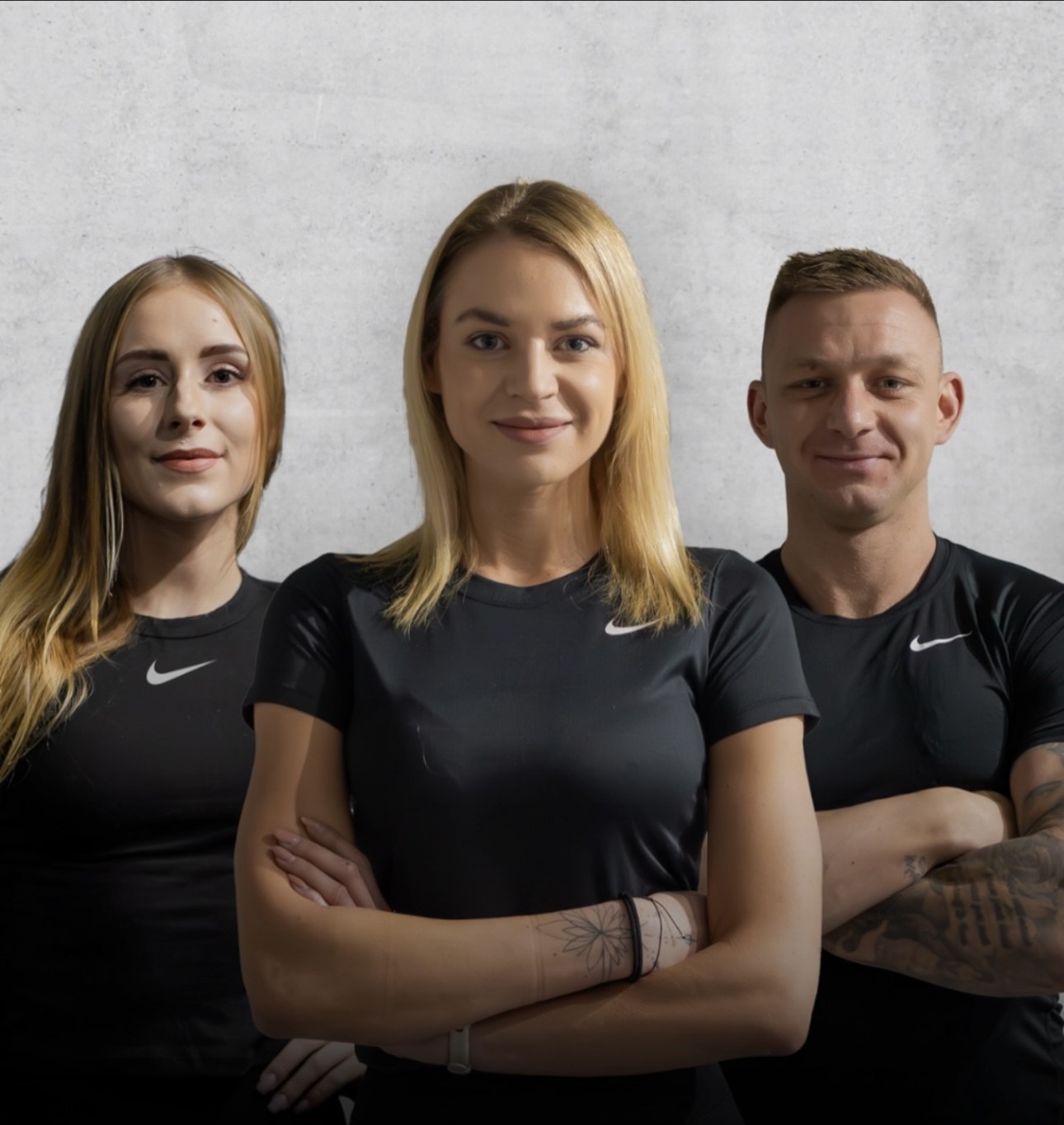 Trzy osoby w czarnych koszulkach Nike stoją na tle szarej ściany, z założonymi rękami, uśmiechnięte, z tatuażami na rękach.