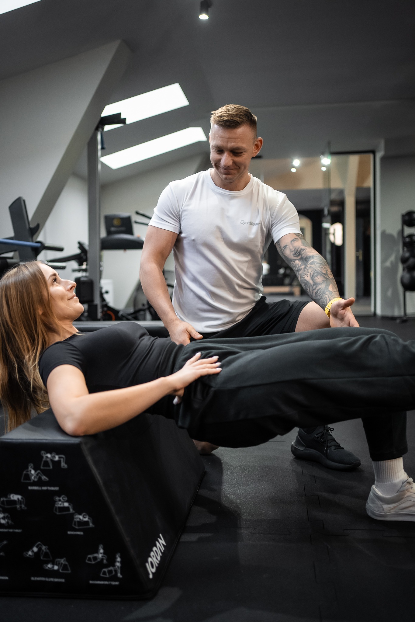 Kobieta wykonuje ćwiczenie bioder na podwyższeniu w obecności mężczyzny, który ją asekuruje, widoczne tatuaże na ramieniu mężczyzny i grafiki z ćwiczeniami na podwyższeniu marki Jordan.