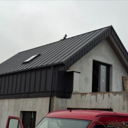 Daniel Dura usługi dekarsko ciesielskie - Nowoczesny dom w trakcie budowy z zamontowanym, ciemnoszarym dachem z blachy na rąbek stojący i oknem dachowym, widoczny fragment czerwonego samochodu dostawczego.