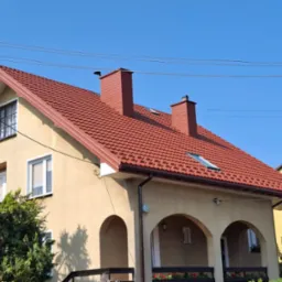 Dom jednorodzinny z nowym, czerwonym dachem z dachówki, dwoma kominami i oknem dachowym, widok z poziomu ogrodu.