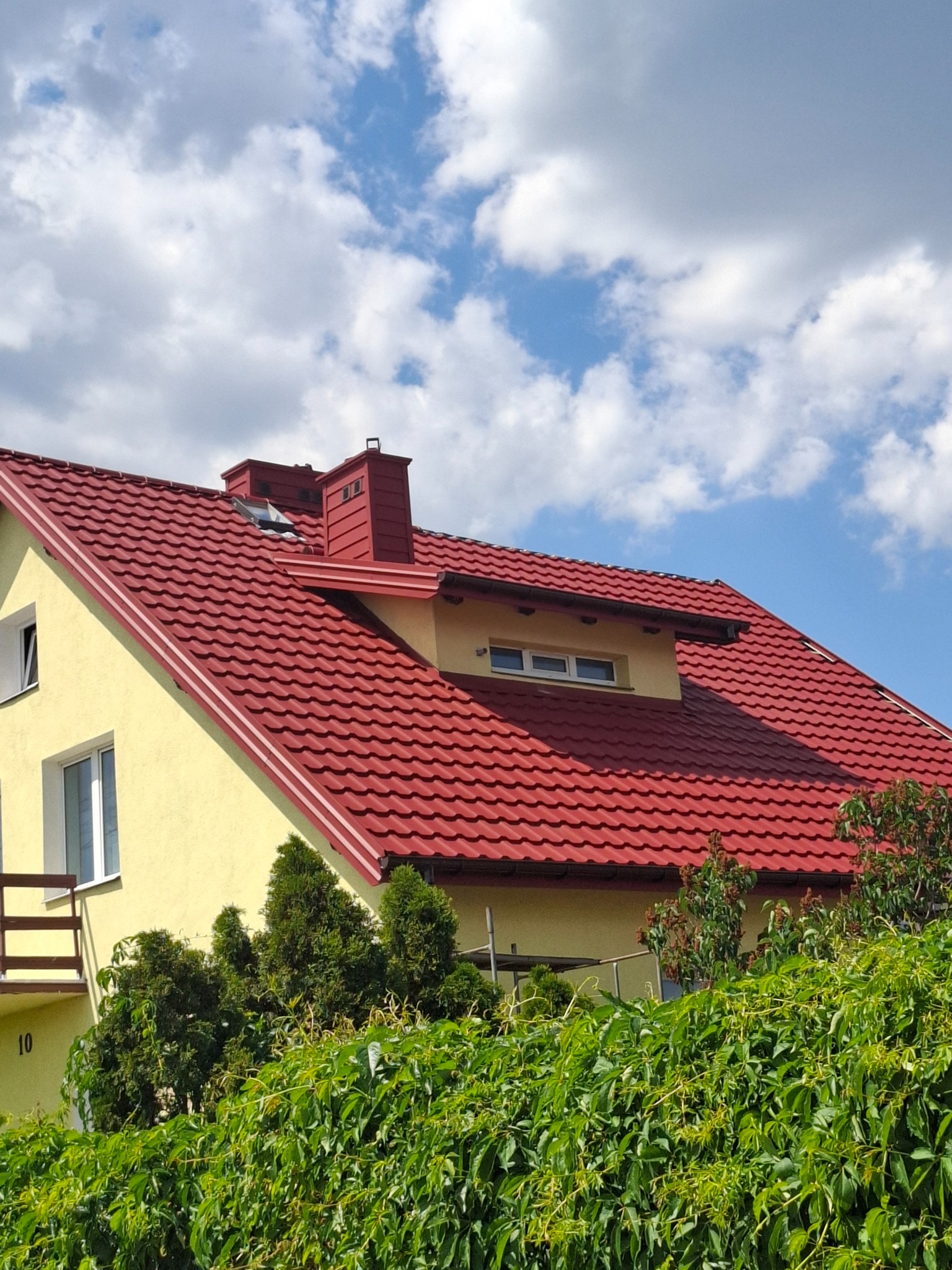 Żółty dom jednorodzinny z nowym, czerwonym dachem z dachówki metalowej, widoczny komin i okno dachowe, na tle błękitnego nieba z białymi chmurami, częściowo zasłonięty zielenią.