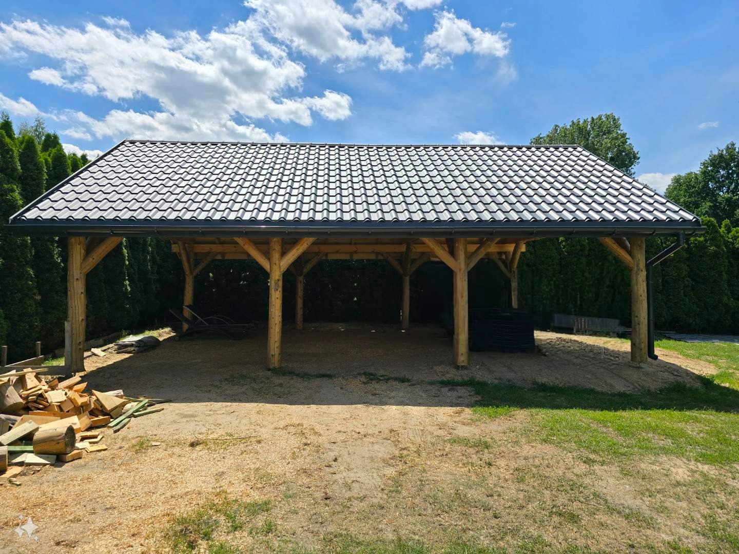 Drewniana wiata z ciemnoszarą dachówką ceramiczną, widoczne rynny, konstrukcja wsparta na drewnianych słupach, w tle iglaki i błękitne niebo z chmurami, na ziemi wióry i resztki drewna po budowie.