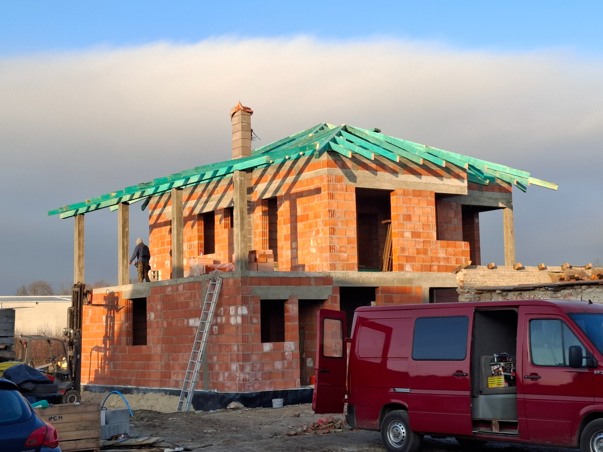 Surowy budynek z czerwonej cegły w trakcie budowy, z widoczną konstrukcją dachu z zielonych łat i kominem. Na budowie obecny pracownik, w tle widać samochód dostawczy z otwartymi drzwiami i wózek...