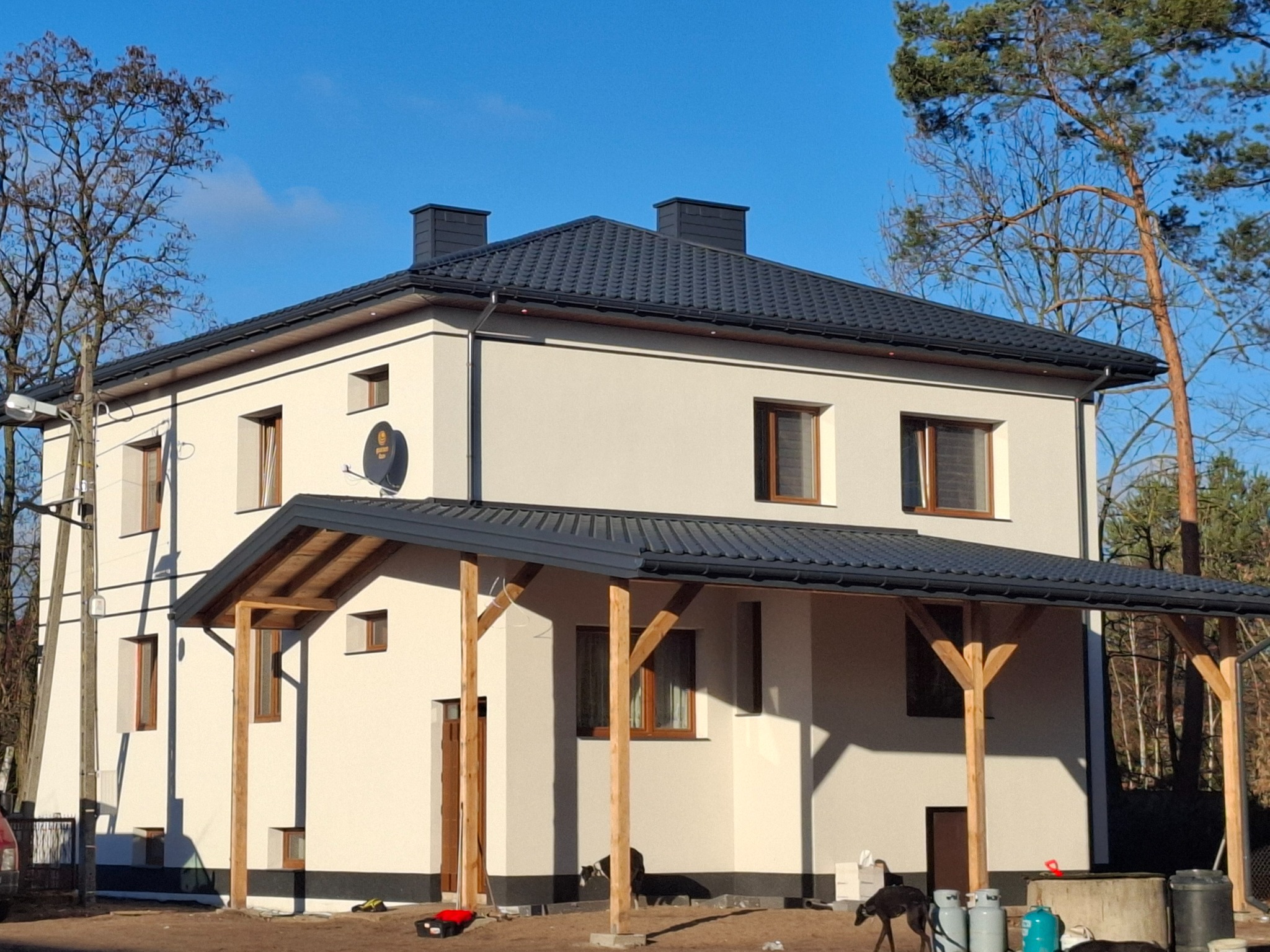 Dwupiętrowy dom z nowym, grafitowym dachem z dachówki, drewnianą wiatą i widocznymi butlami gazowymi przy fundamencie.