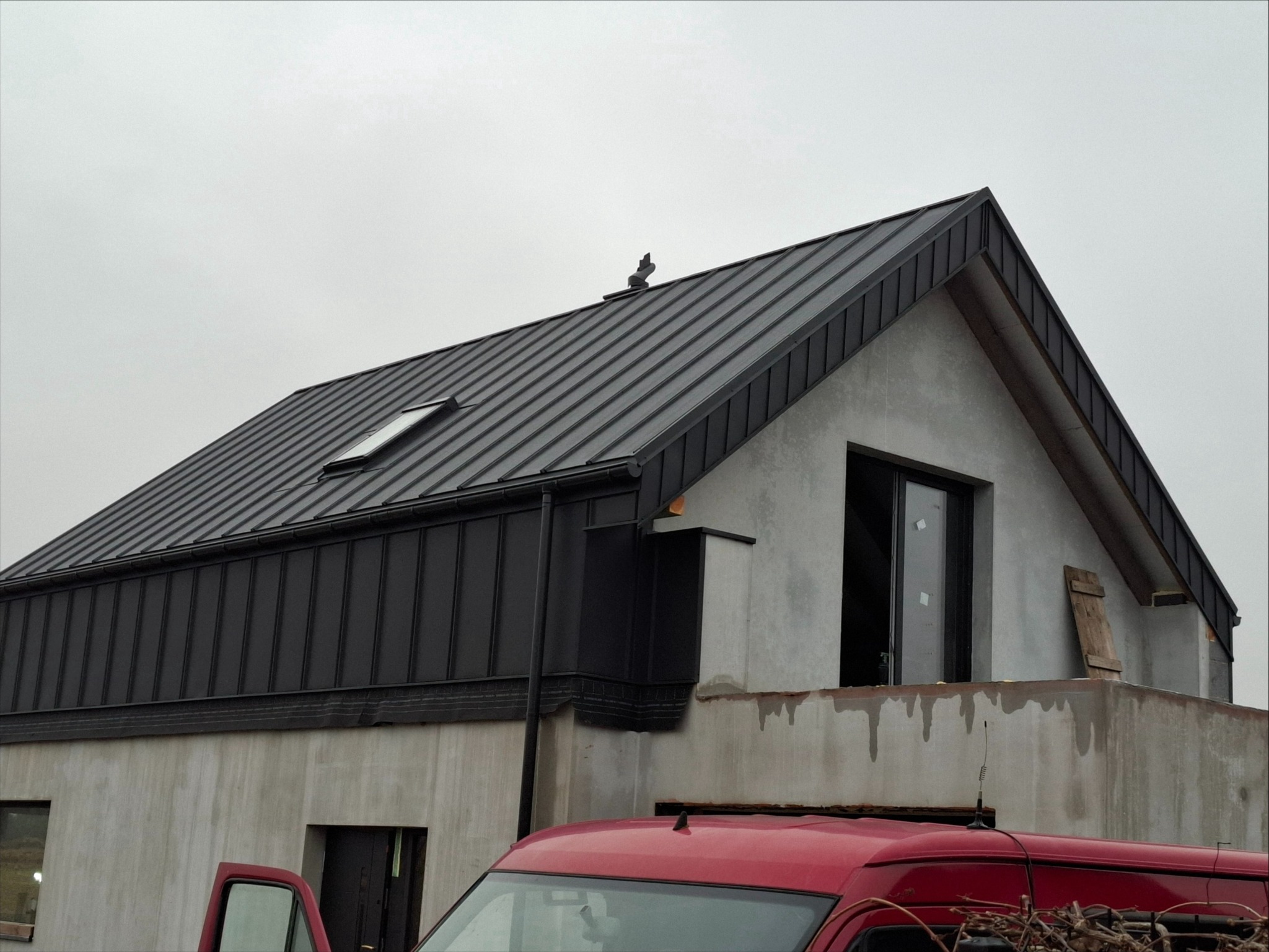 Nowoczesny dom w trakcie budowy z zamontowanym, ciemnoszarym dachem z blachy na rąbek stojący i oknem dachowym, widoczny fragment czerwonego samochodu dostawczego.