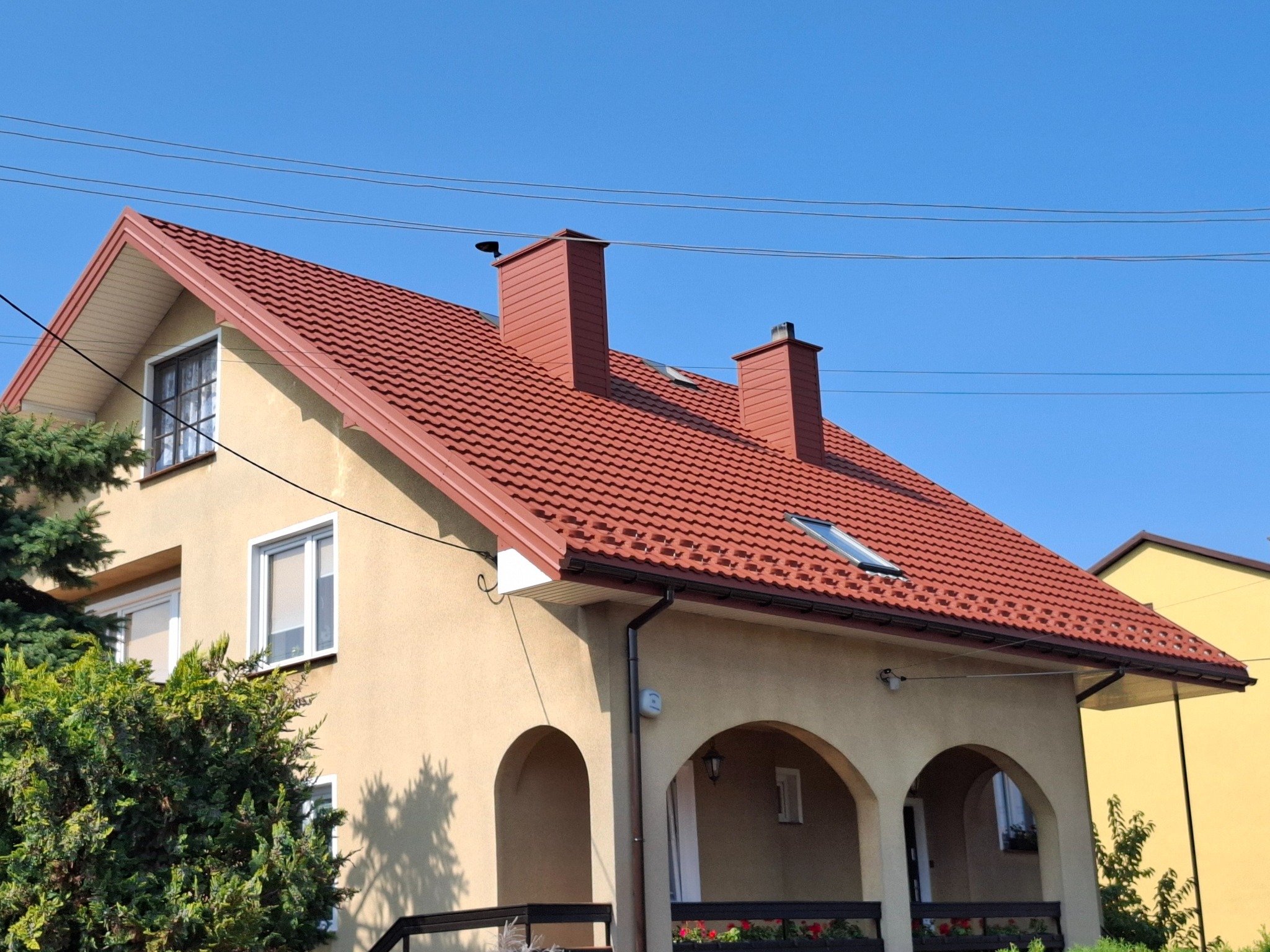 Dom jednorodzinny z nowym, czerwonym dachem z dachówki, dwoma kominami i oknem dachowym, widok z poziomu ogrodu.