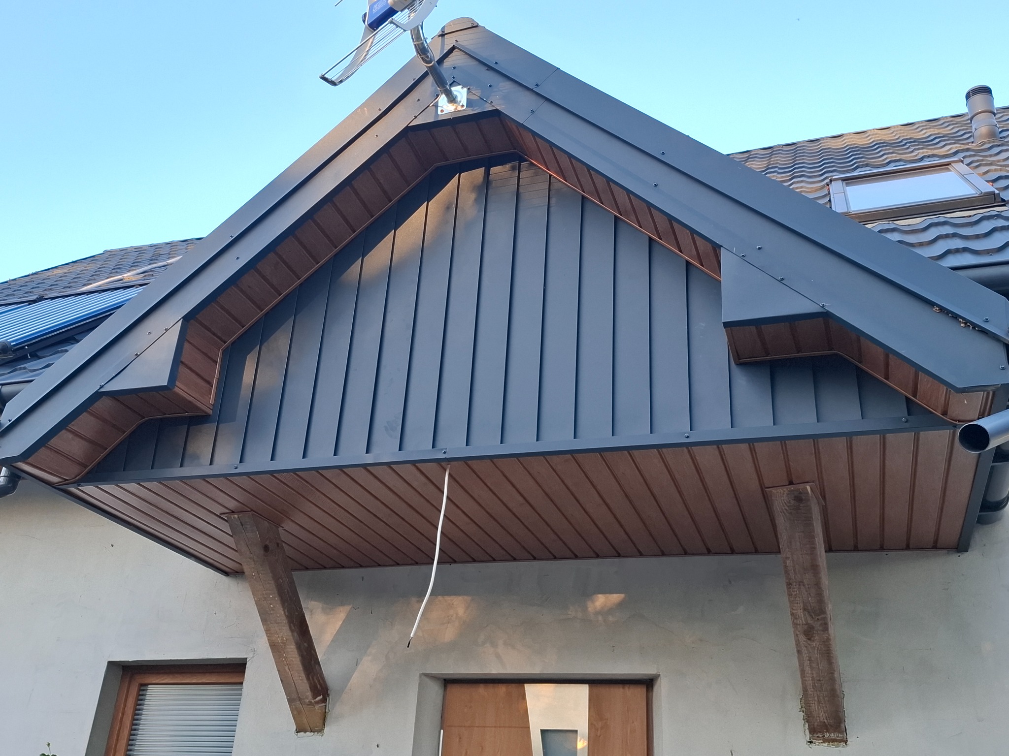 Zadaszenie nad wejściem do domu w Lipcach Reymontowskich, z ciemnoszarą blachą i drewnianą podbitką, wsparte na drewnianych belkach, widoczne okno dachowe i antena.