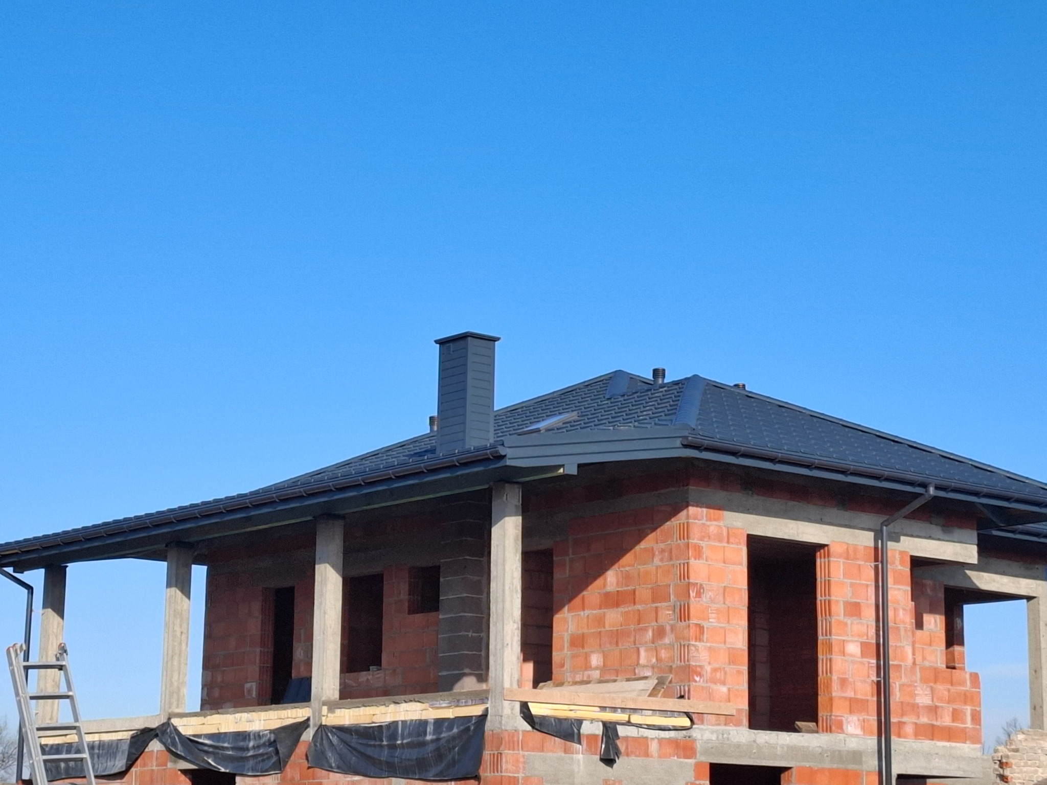 Niedokończony budynek z nowym, ciemnoszarym dachem z dachówki i kominem, widoczne rynny, ceglana konstrukcja w stanie surowym, drabina oparta o budynek.