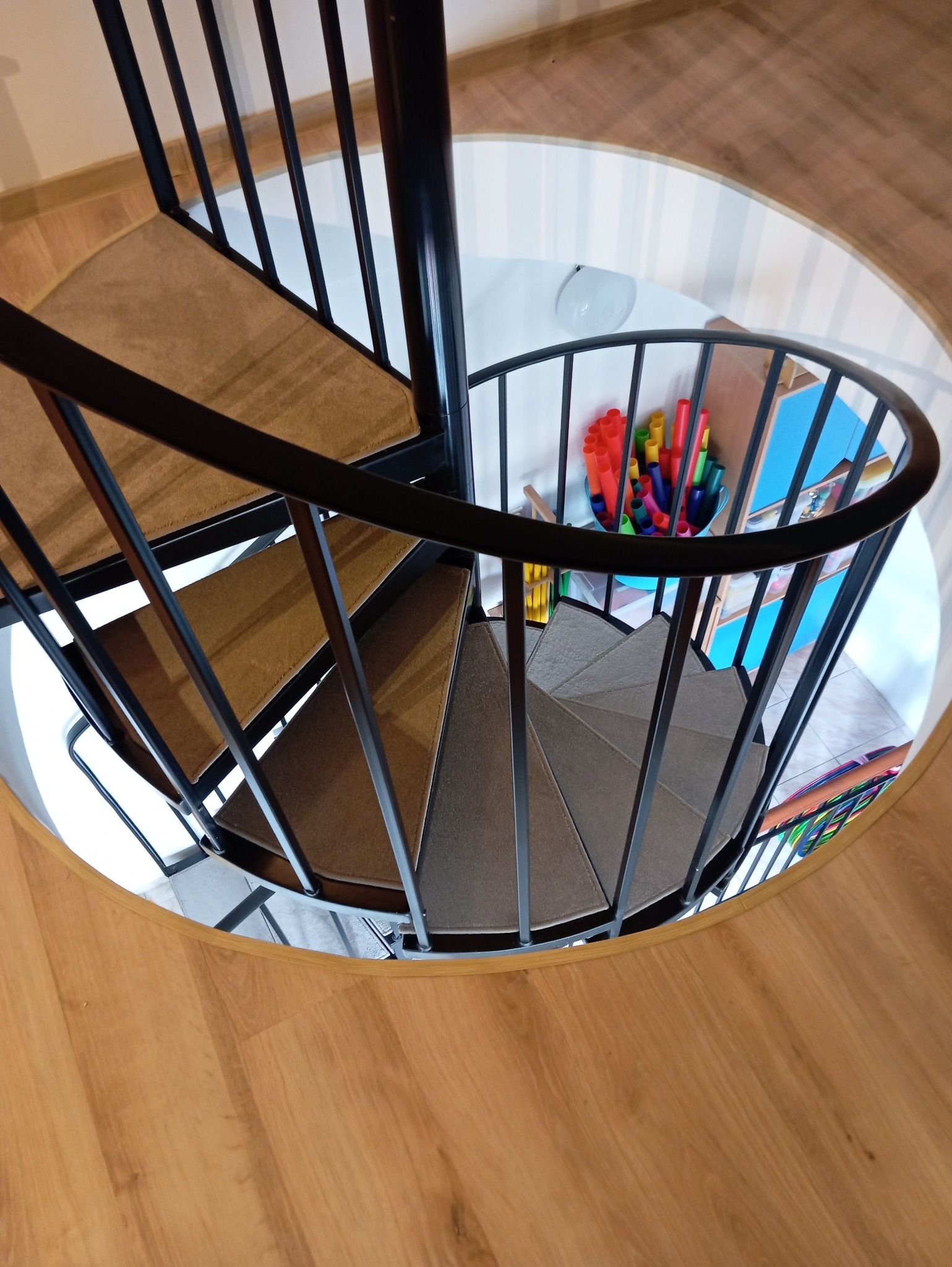 Widok z góry na spiralne schody z czarną metalową balustradą i stopniami pokrytymi brązową wykładziną dywanową, otoczone drewnianą podłogą, z widocznym fragmentem pokoju dziecięcego w tle.